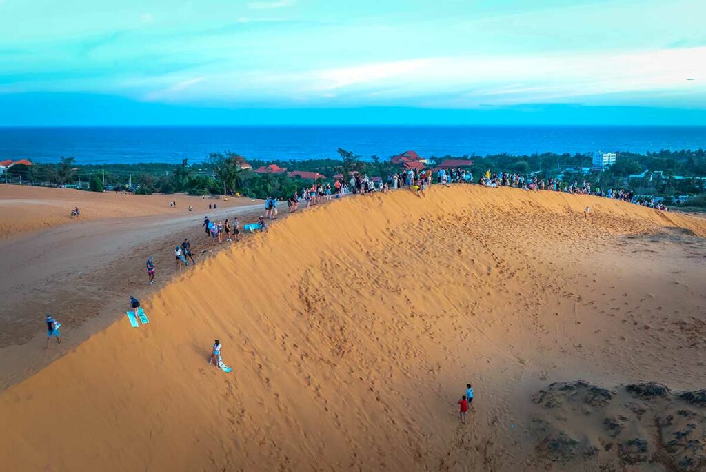 Luchtfoto van drukke rode zandduinen van Mui Ne bij zonsondergang met tientallen bezoekers die met sleetjes naar beneden glijden en de zee op de achtergrond — de duinen zijn populair maar kunnen erg druk zijn
