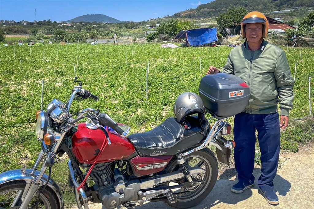 Easy Rider-gids staat naast zijn rode motor op het platteland buiten Dalat, met groene groentevelden op de achtergrond — bezoeken aan lokale boerderijen zijn vaste stops op een Easy Rider tour