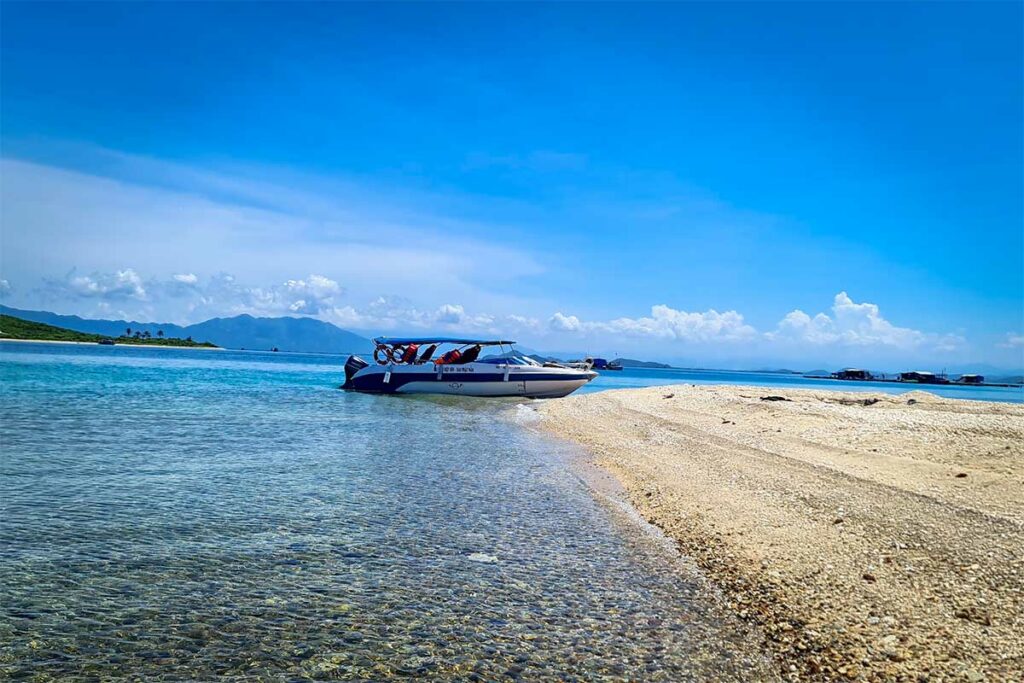 Speedboot aangemeerd op een smal zandbankie in de Van Phong Bay met helder ondiep water en eilanden zichtbaar op de achtergrond — speedboottours verbinden de eilanden in de baai