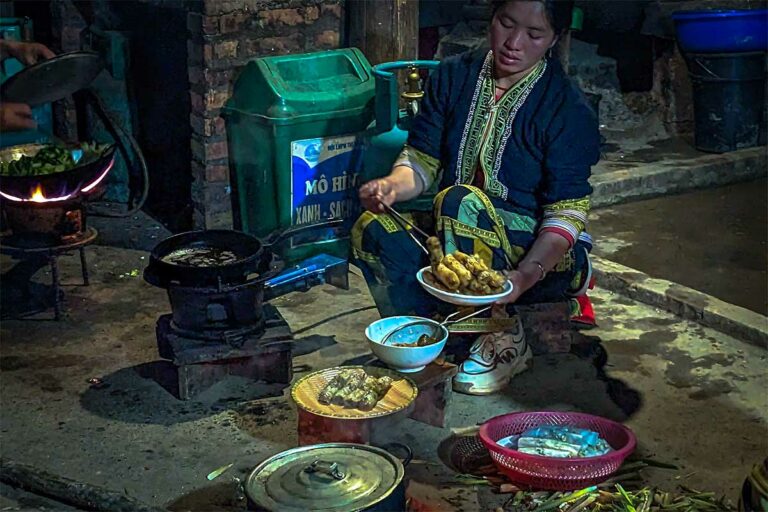 Vrouw in traditionele kledij van een etnische minderheid bereidt meerdere gerechten tegelijk in de keuken van een homestay in Sapa
