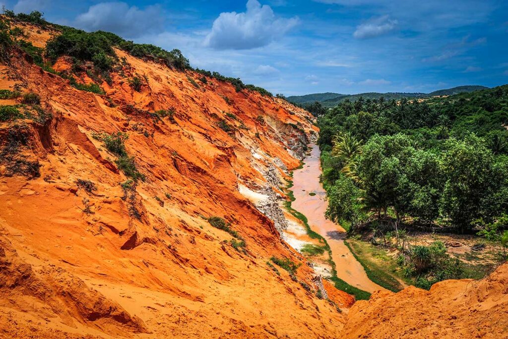 Bovenaanzicht van de rode canyon van de Fairy Stream in Mui Ne met het slingerende beekje en palmbomen op de achtergrond