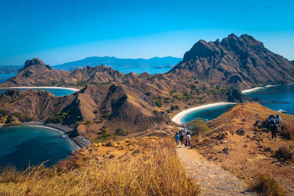 Groep wandelaars op een steenpad over de dorre heuvels van Padar Island bij Flores in Indonesie, met drie baaien en witte stranden zichtbaar op de achtergrond