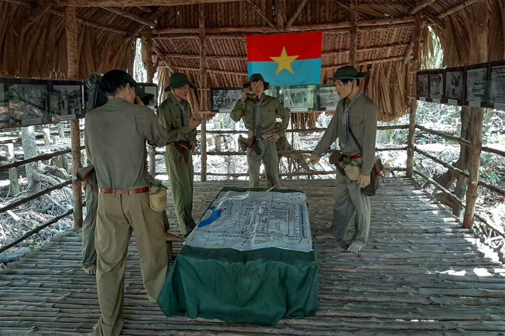 Wassenbeelden van Vietcong-commandanten die een militaire kaart bestuderen in een rieten schuilhut in het Sac bos van het Can Gio reservaat, met de vlag van het Nationaal Bevrijdingsfront aan de wand — de guerrillabasis in het Sac bos speelde een sleutelrol tijdens de Vietnamoorlog