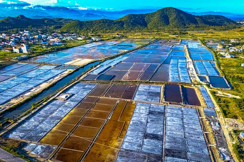 Luchtfoto van de uitgestrekte Hon Khoi zoutvelden met tientallen rechthoekige zoutpannen in verschillende kleuren, een dorp aan de rand en groene heuvels op de achtergrond bij Doc Let