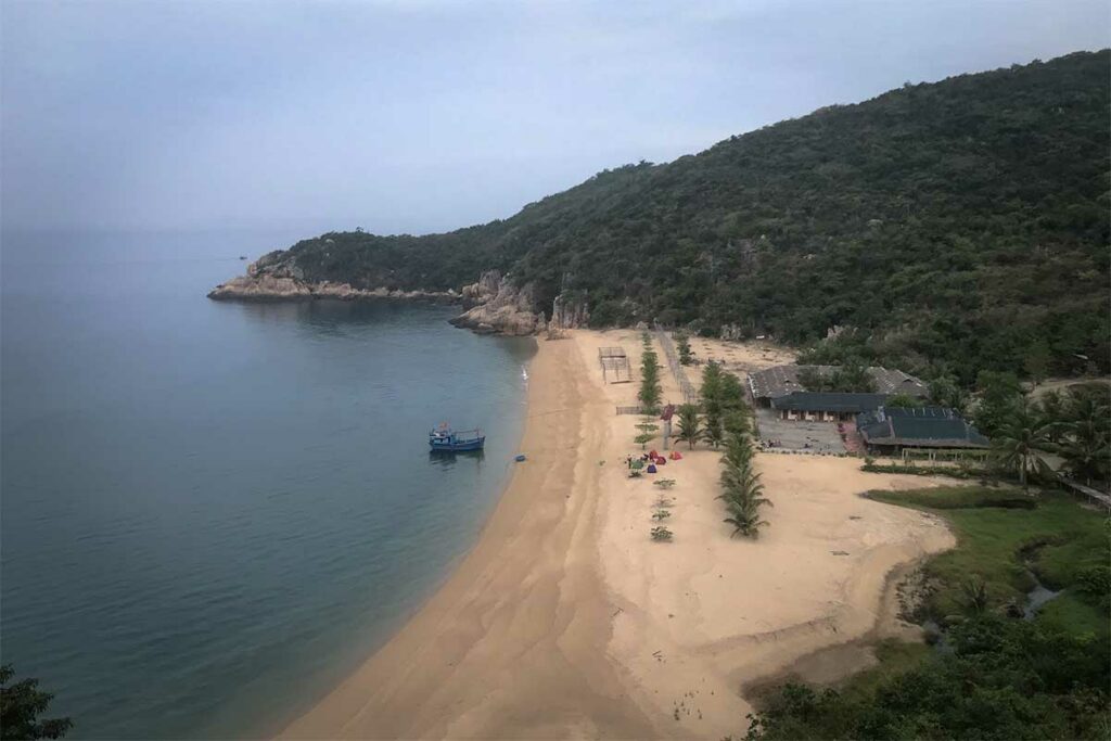 Verlaten goudgeel strand op Hon Lon eiland in de Van Phong Bay met een vissersboot in het rustige water en beboste heuvels op de achtergrond — een van de weinig ontwikkelde eilanden in de baai