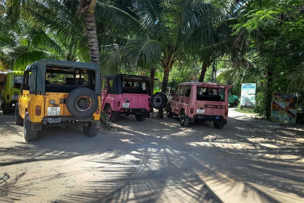 Gele en roze jeeps geparkeerd onder palmbomen bij de ingang van de Fairy Stream in Mui Ne — jeeptours combineren vaak de duinen met een bezoek aan de Fairy Stream