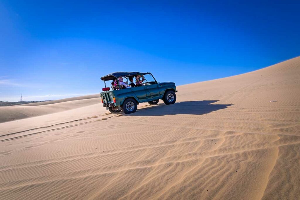 Groene jeep rijdt met toeristen over de witte zandduinen van Mui Ne onder een strakblauwe lucht — jeeptours zijn een populaire manier om de duinen comfortabel te verkennen