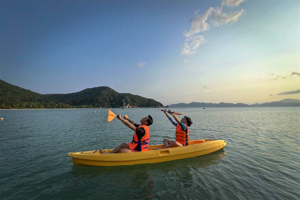 Twee kajakkers in oranje reddingsvesten heffen hun peddels omhoog in een geel kajak op het rustige water van Ninh Van Bay bij zonsondergang, met beboste heuvels en eilanden op de achtergrond