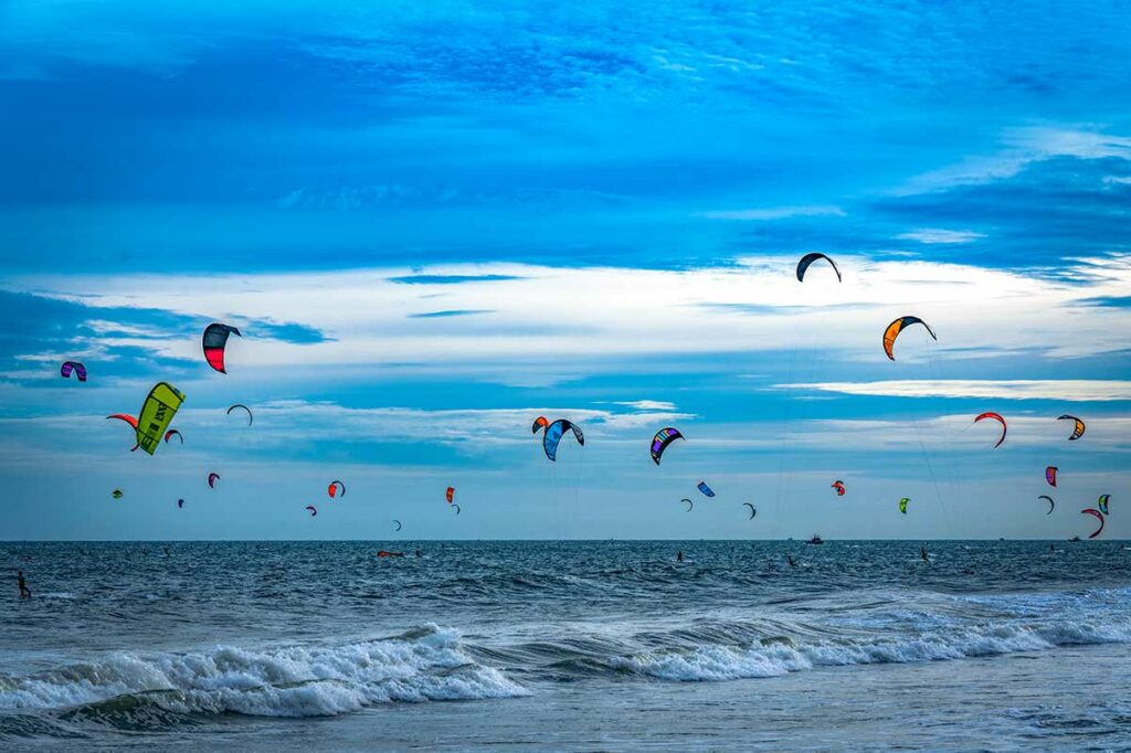 Tientallen kleurrijke kitevliegers in de lucht boven de zee van Mui Ne bij bewolkte hemel — een typisch beeld tijdens het kitesurfseizoen in Mui Ne
