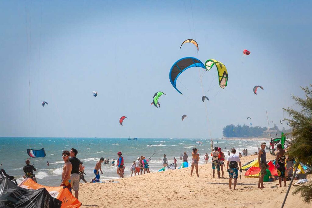 Druk strand in Mui Ne met kitesurfers, toeschouwers en vliegers boven de zee — het strand staat bekend als een van de populairste kitesurflocaties van Zuidoost-Azië