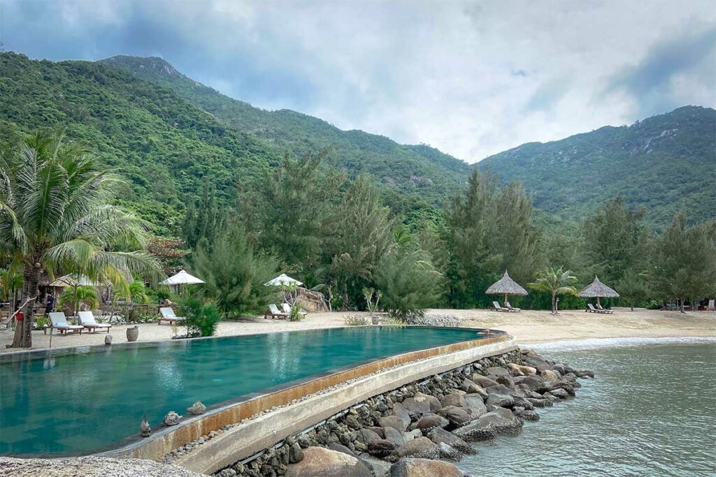 Zwembad van het Lalya resort aan de rand van het strand in Ninh Van Bay, omgeven door dennenbomen en groene berghellingen — een van de exclusieve resorts die alleen per boot bereikbaar zijn