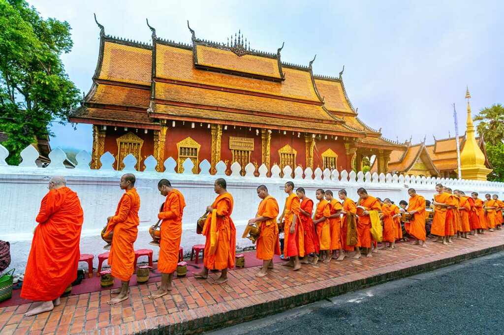 Rij boeddhistische monniken in oranje gewaden tijdens de dagelijkse aalmoezentocht langs een gouden tempel in Luang Prabang, Laos