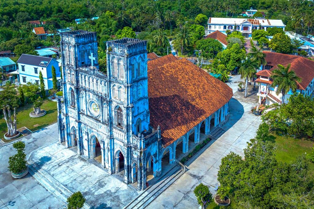 Luchtfoto van de Mang Lang kerk in Phu Yen, een negentiende-eeuwse Franse gotische kerk met blauwe gevels en rode dakpannen, omgeven door palmbomen en een dorpje — een van de oudste kerken in centraal Vietnam