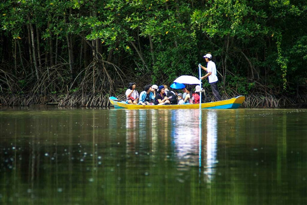 Groep bezoekers in een gele houten boot tijdens een boottocht door de mangrovekreken van het Can Gio reservaat, begeleid door een staande gids met een pols — de boottocht voert langs dichte mangroveoevers met luchtwortels en spiegelend water