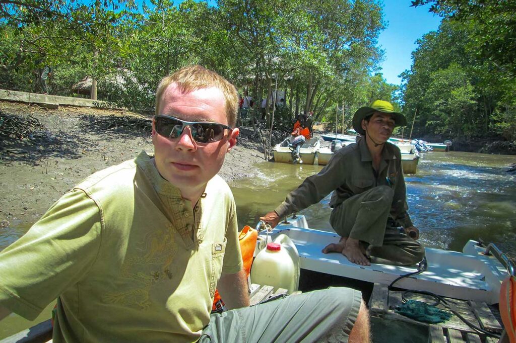 Toerist en lokale gids op een motorboot tijdens een boottocht door het Can Gio reservaat, met mangrovebomen langs de modderige oever op de achtergrond — een boottocht door het reservaat is de meest gebruikelijke manier om het mangrovegebos te verkennen