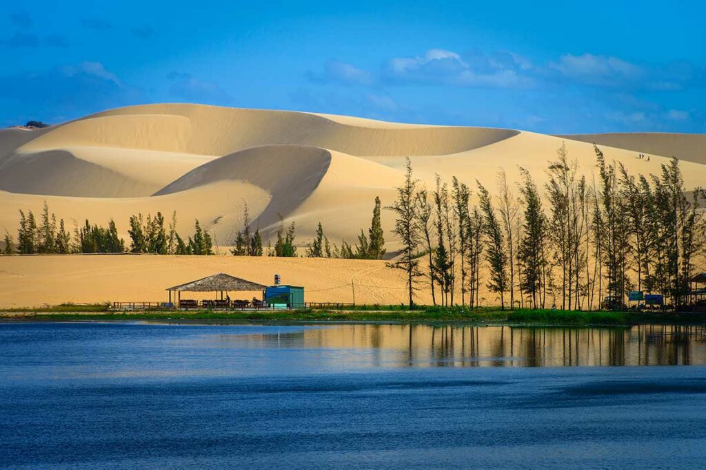 Meer met weerspiegeling van de witte zandduinen van Mui Ne, omzoomd door dennenbomen en een kleine paviljoenstent op de oever — het meer aan de voet van de duinen is een minder bekend maar mooi onderdeel van het landschap