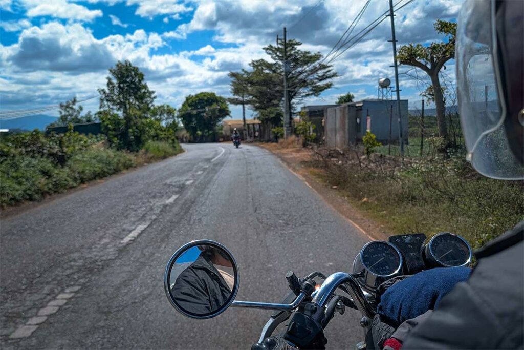Uitzicht vanuit het zadel van een Easy Rider-motor op een rechte weg door het platteland bij Dalat, met een andere motor voorop en bewolkte lucht boven de vlakte — een typisch beeld tijdens een dagrit vanuit Dalat