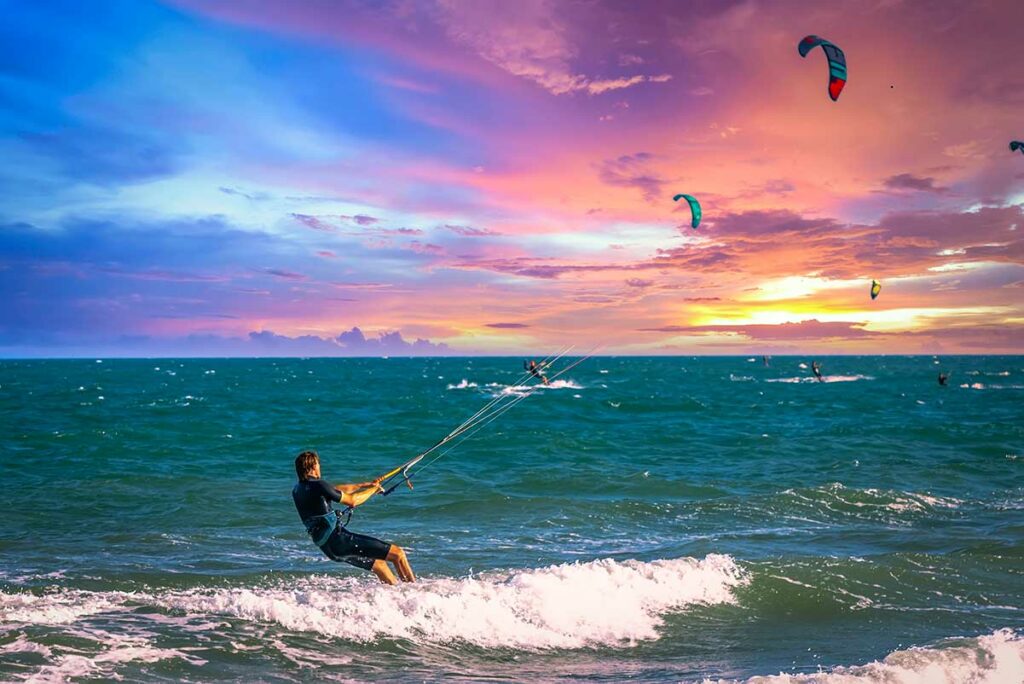 Kitesurfer scheert door de golven bij zonsondergang in Mui Ne met andere surfers en vliegers zichtbaar op de achtergrond — de constante wind maakt Mui Ne ideaal voor kitesurfen