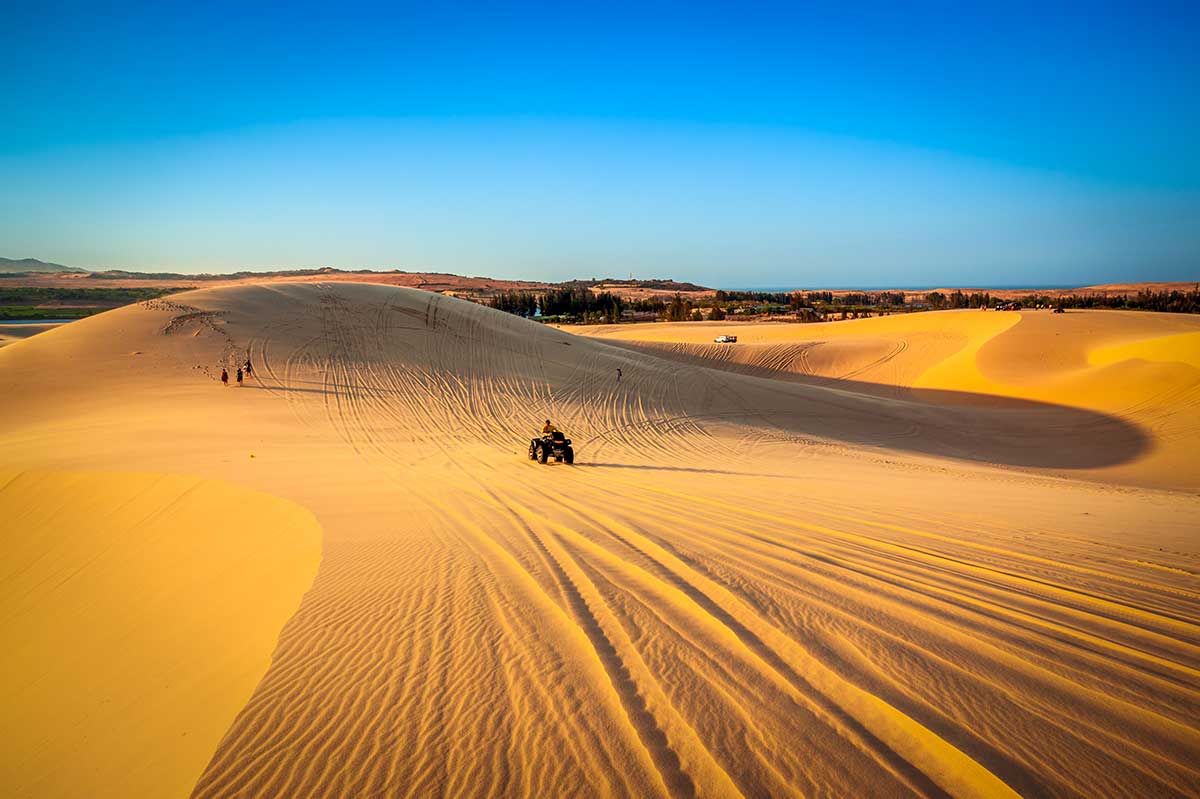 Quad rijdt over de goudgele zandduinen van Mui Ne bij laagstaande zon met sporen in het zand en de kustlijn op de achtergrond — een overzichtsfoto die het uitgestrekte duinenlandschap van Mui Ne goed weergeeft