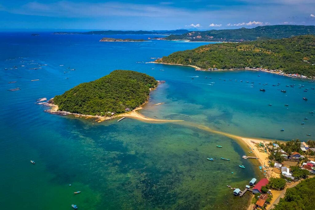 Luchtfoto van het eiland Nhat Tu Son in Phu Yen met een beboste heuvel, een zandbank die het eiland verbindt met de kust en tientallen visserboten in het turquoise water