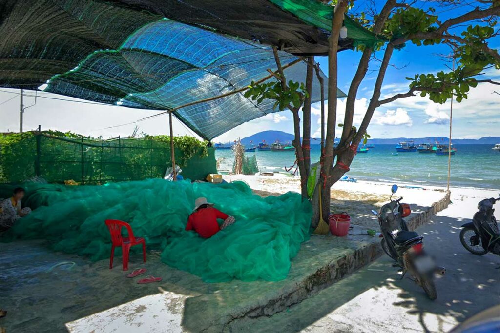Vrouw repareert een groot groen visnet in het vissersdorp Ninh Thuy bij Doc Let Beach, met visserboten zichtbaar op het water op de achtergrond — een alledaags tafereel in deze actieve vissersgemeenschap