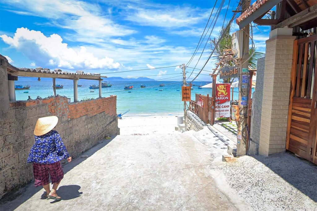 Smalle betonnen steeg in het vissersdorp Ninh Thuy met zicht op het turquoise water van Doc Let Beach, visserboten in de baai en lokale horecabordjes langs de kant