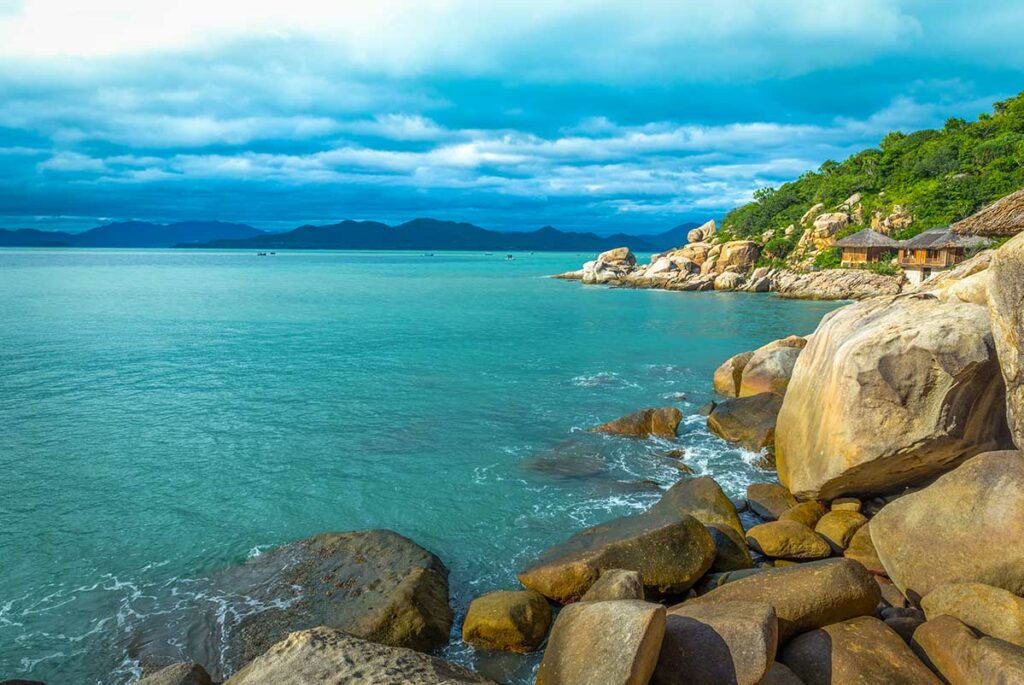 Rotsachtige kustlijn van Ninh Van Bay met granieten keien, turquoise water en rieten bungalows van een luxeresort verscholen tussen de beboste heuvels op de achtergrond