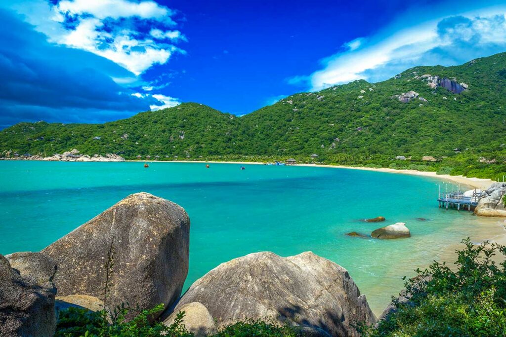 Panoramisch uitzicht op de Ninh Van Bay met granieten rotsblokken op de voorgrond, turquoise water, een wit zandstrand en rieten bungalows van een luxeresort langs de kustlijn