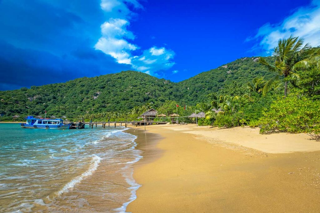Breed goudgeel strand van Ninh Van Bay met witte golven, palmbomen en een houten pier in de verte — het strand is onderdeel van een exclusief resort dat niet openbaar toegankelijk is