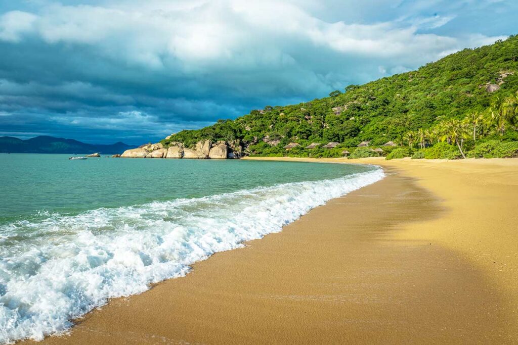 Breed goudgeel strand van Ninh Van Bay met opkomende golven, palmbomen en rieten bungalows zichtbaar in de jungle — een van de mooiste en meest afgelegen baaien bij Nha Trang