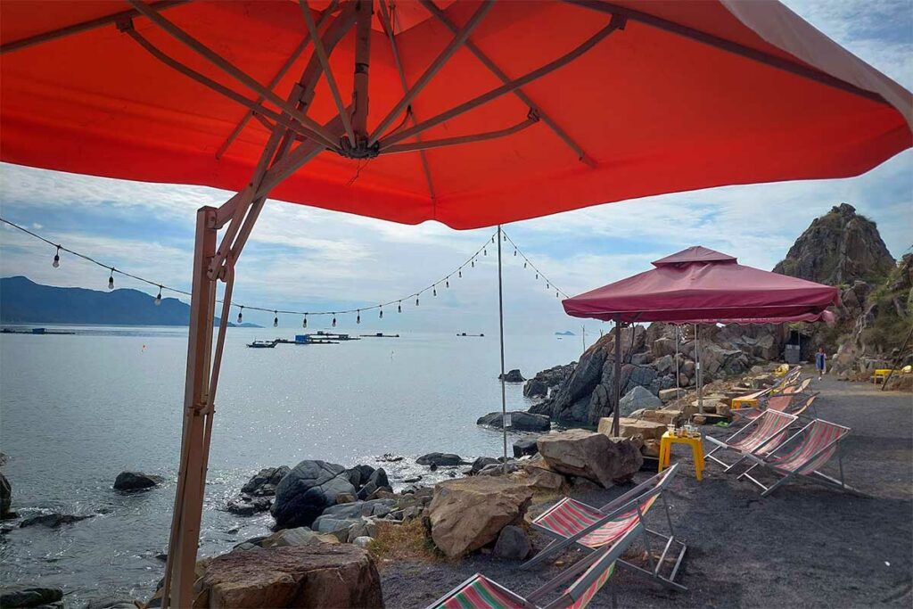 Uitzichtcafe langs de rotskust van Ninh Van Bay met rode parasols, ligbedden en een panoramisch zeezicht op de baai — een informele plek om de omgeving te verkennen