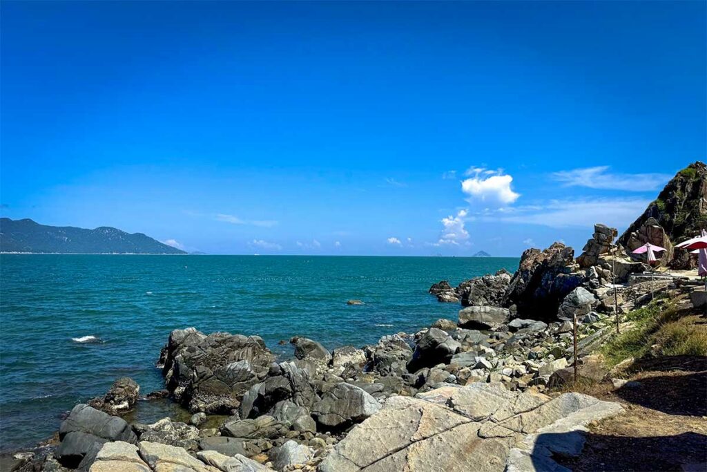 Rotskust van Ninh Van Bay met grote donkere keien, turquoise water en een klein cafe met parasols op de achtergrond — de ruige kustlijn maakt Ninh Van Bay uniek langs de kust van Nha Trang
