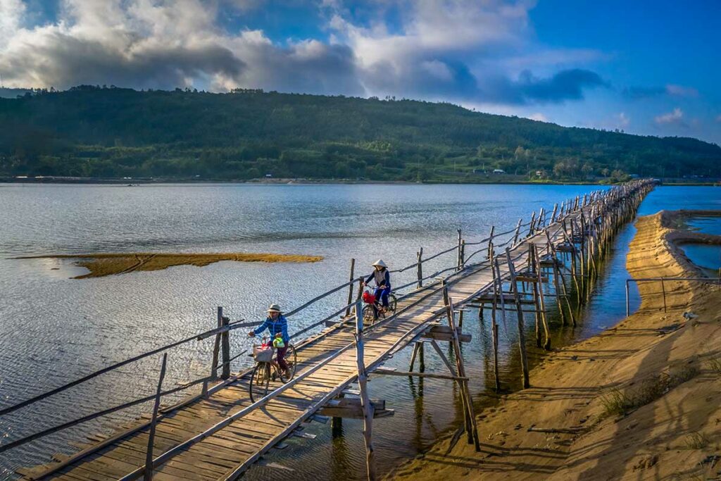 Twee fietsers met Vietnamese hoeden rijden over de smalle houten Ong Cop brug in Phu Yen, met een brede lagune en beboste heuvel op de achtergrond — een van de langste houten bruggen van Vietnam