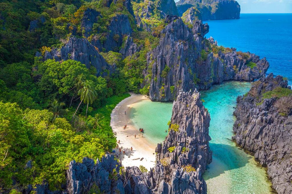 Luchtfoto van een afgelegen wit zandstrand ingesloten door scherpe kalkstenen rotswanden en turquoise water op Palawan in de Filipijnen, met een handvol bezoekers op het strand