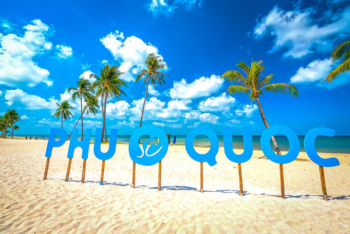 Groot blauw Phu Quoc-bord op een wit zandstrand met palmbomen en een strakblauwe lucht — het droge seizoen van november tot april is de beste reistijd voor Phu Quoc