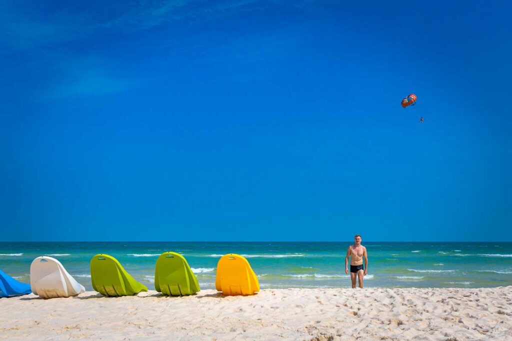 Toerist staat op het witte strand van Phu Quoc bij heldere blauwe lucht, met kleurrijke watersportmaterialen op het zand en een parasailer in de lucht — watersport is mogelijk tijdens het droge seizoen
