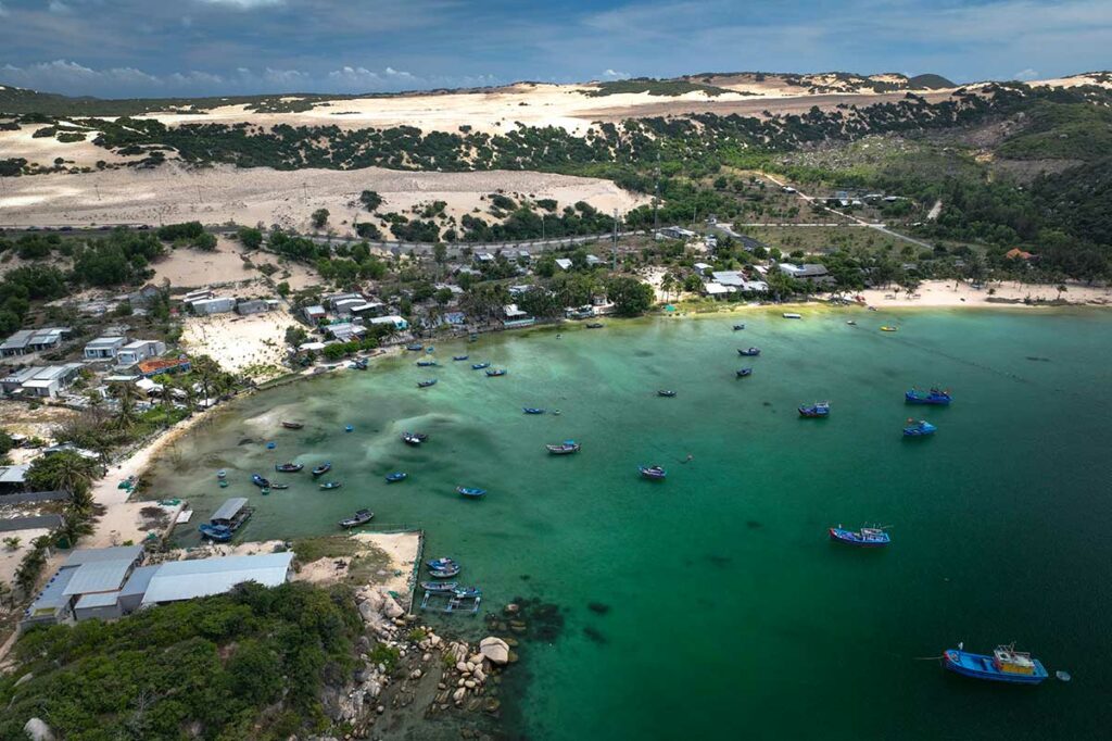 Luchtfoto van de vissershaven van Dam Mon met tientallen kleurrijke boten op het groene water, een klein vissersdorp en zandduinen op de achtergrond in de Van Phong Bay