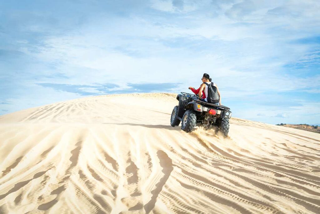 Quad rijdt met twee passagiers over de witte zandduinen van Mui Ne met een blauwe lucht op de achtergrond — een actieve manier om de duinen van Mui Ne te verkennen