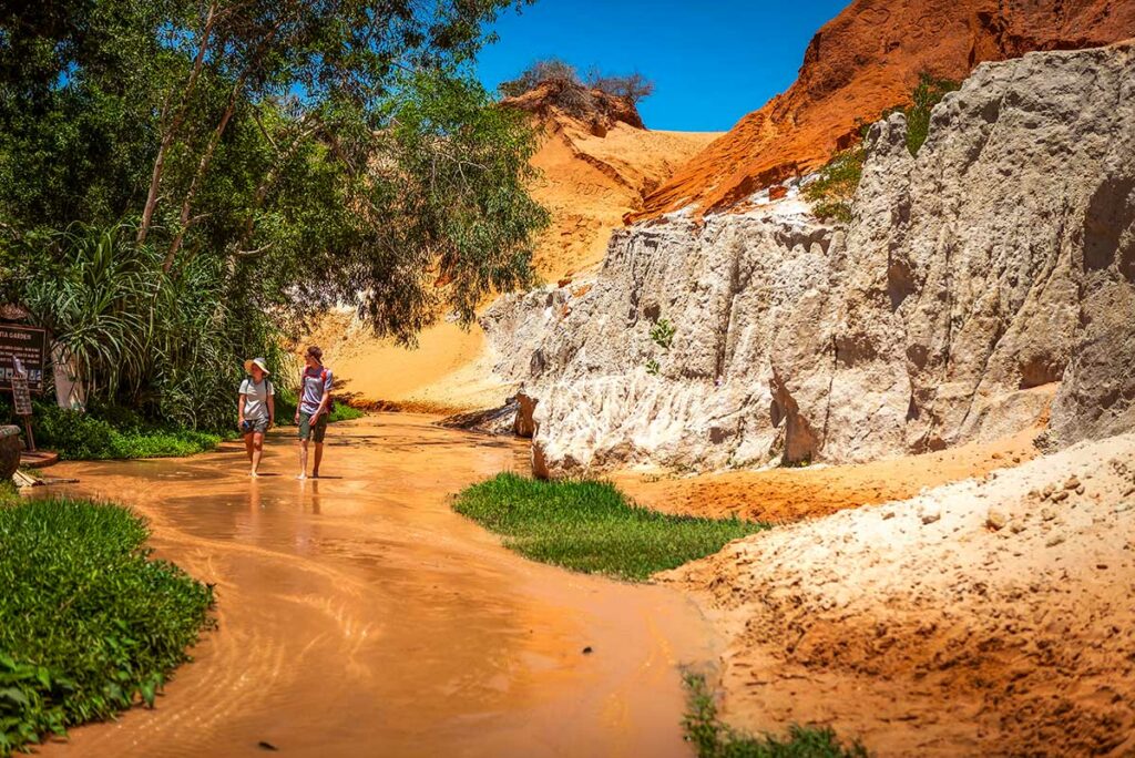 Twee reizigers lopen barrevoets door het ondiepe water van de Fairy Stream in Mui Ne langs witte en rode zandsteenwanden — schoenen uitdoen is vereist voor dit wandelpad