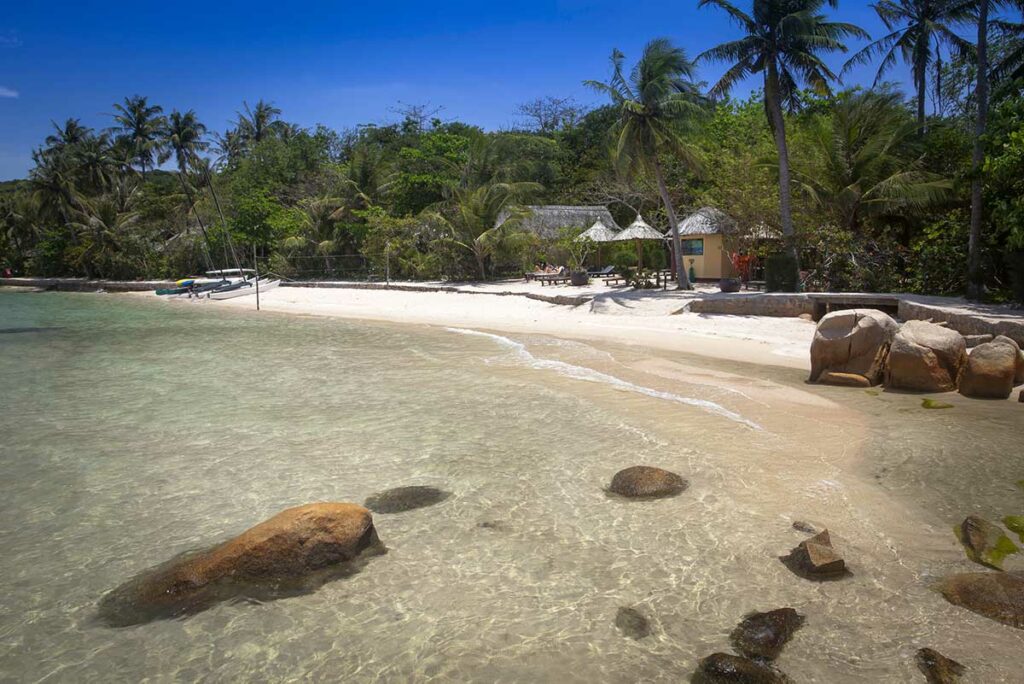 Rustig wit strand van Whale Island met rieten strandhuisjes, palmbomen en helder turquoise water — een van de meest afgelegen resortbestemmingen in de Van Phong Bay