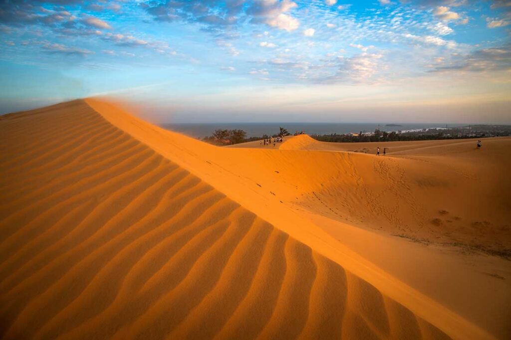 Golvende rode zandduinen van Mui Ne in het warme avondlicht met kleine figuren van bezoekers op de top — de rode duinen zijn het beste te bezoeken bij zonsondergang