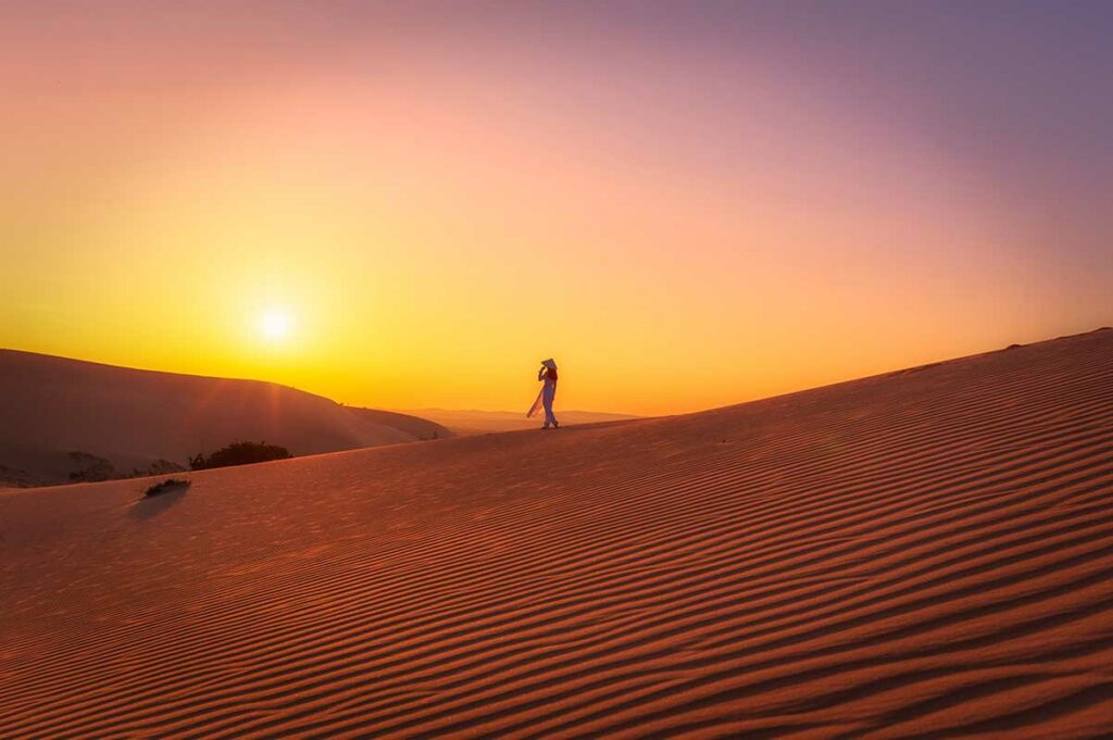 Silhouet van een vrouw met conische hoed op de rode zandduinen van Mui Ne bij zonsondergang met een paarsroze lucht — zonsondergang op de duinen is een van de mooiste momenten van een bezoek aan Mui Ne