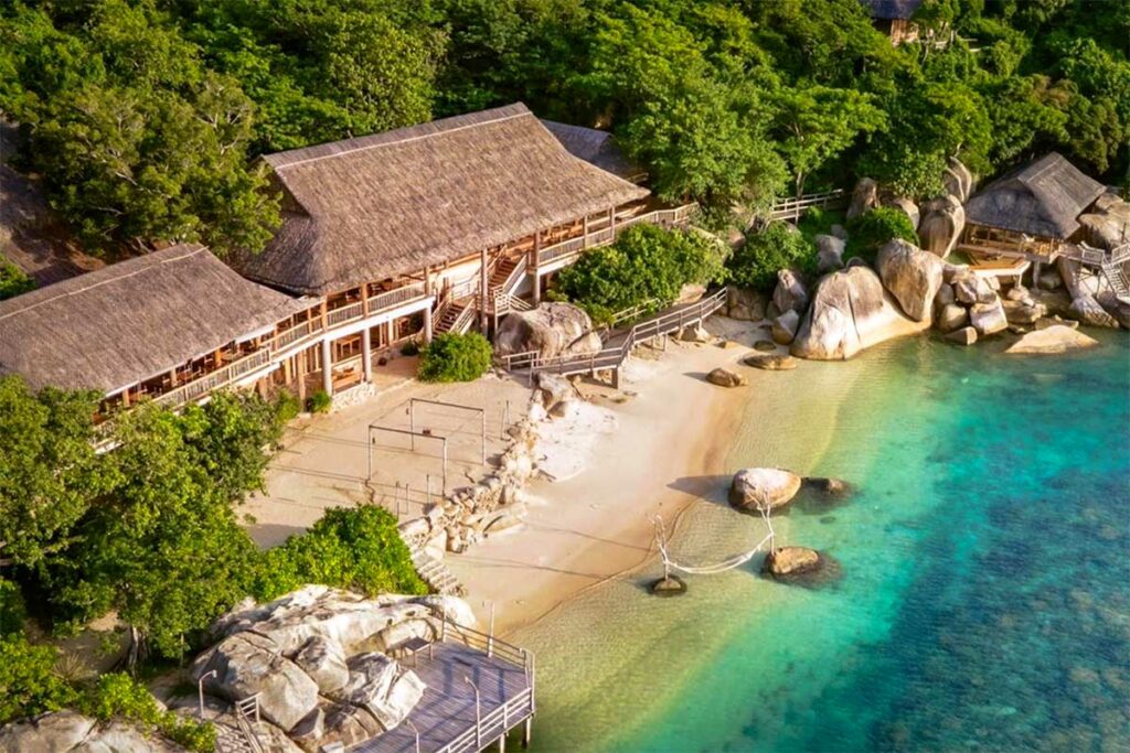 Luchtfoto van het Six Senses resort in Ninh Van Bay met rieten bungalows verscholen tussen rotsen en jungle, een zwembad direct aan het water en een houten pier — bereikbaar alleen per boot