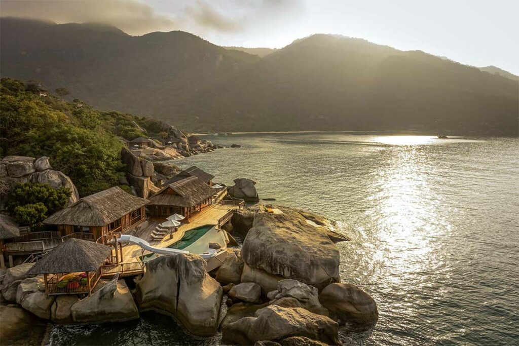 Luchtfoto van het Six Senses resort langs de rotsachtige kustlijn van Ninh Van Bay bij avondlicht, met een overloopzwembad, houten terrassen en bungalows verspreid over de rotsen aan het water