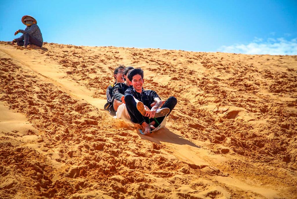 Lachende jongeren glijden op een slee van plastic naar beneden over de rode zandduinen van Mui Ne — sandboarden op de duinen is een goedkope activiteit die je ter plekke kunt huren