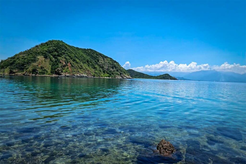Kristalhelder water in de Van Phong Bay met zichtbare stenen op de bodem en een beboste heuvel op de achtergrond — het heldere water maakt snorkelen in de Van Phong Bay erg aantrekkelijk