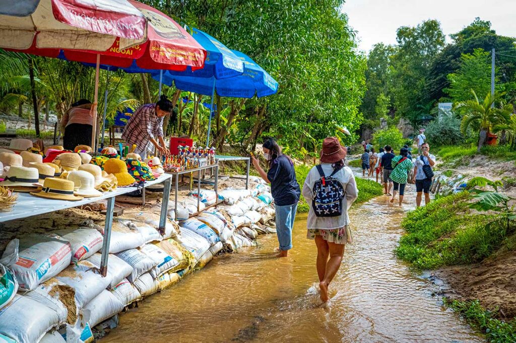 Toeristen lopen barrevoets langs souvenirstalletjes met hoeden en snuisterijen in het beekje bij de ingang van de Fairy Stream in Mui Ne — de commerciële sfeer aan het begin van het parcours