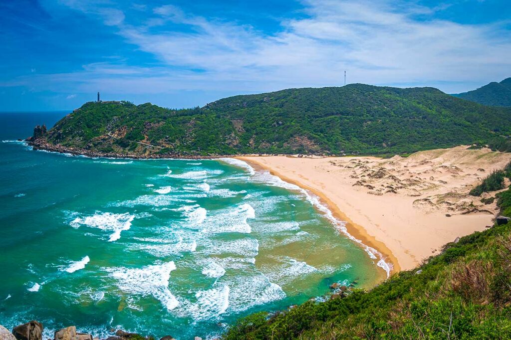Verlaten zandstrand van Bai Mon Beach bij Mui Dien in Phu Yen met krachtige golven, zandduinen en een vuurtoren zichtbaar op de beboste kaap — het meest oostelijke punt van het Vietnamese vasteland op het vasteland