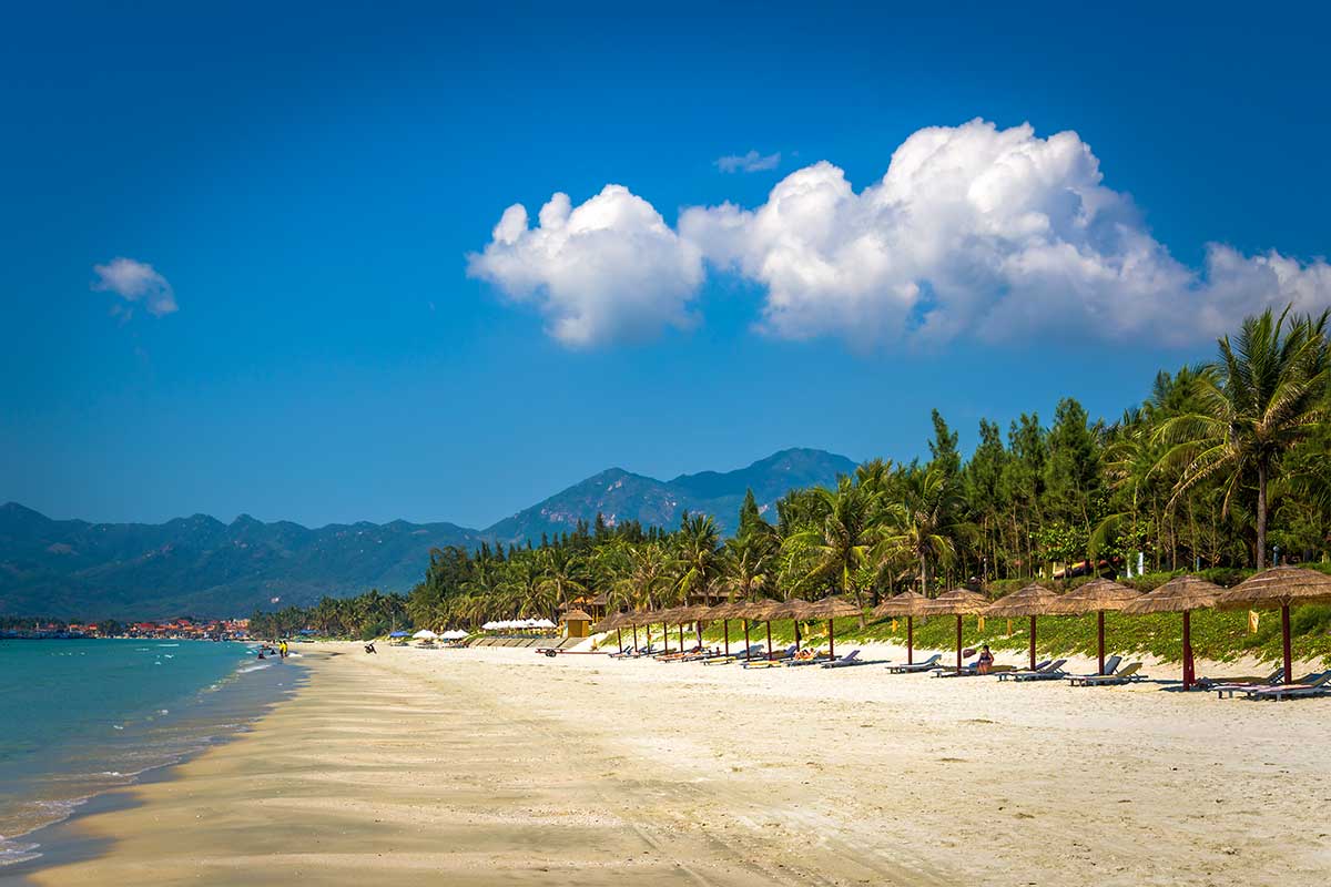 Lang wit strand van Doc Let Beach met rieten parasols, ligbedden, palmbomen en bergen op de achtergrond — het strand ligt op etwa een uur rijden van Nha Trang en trekt relatief weinig toeristen