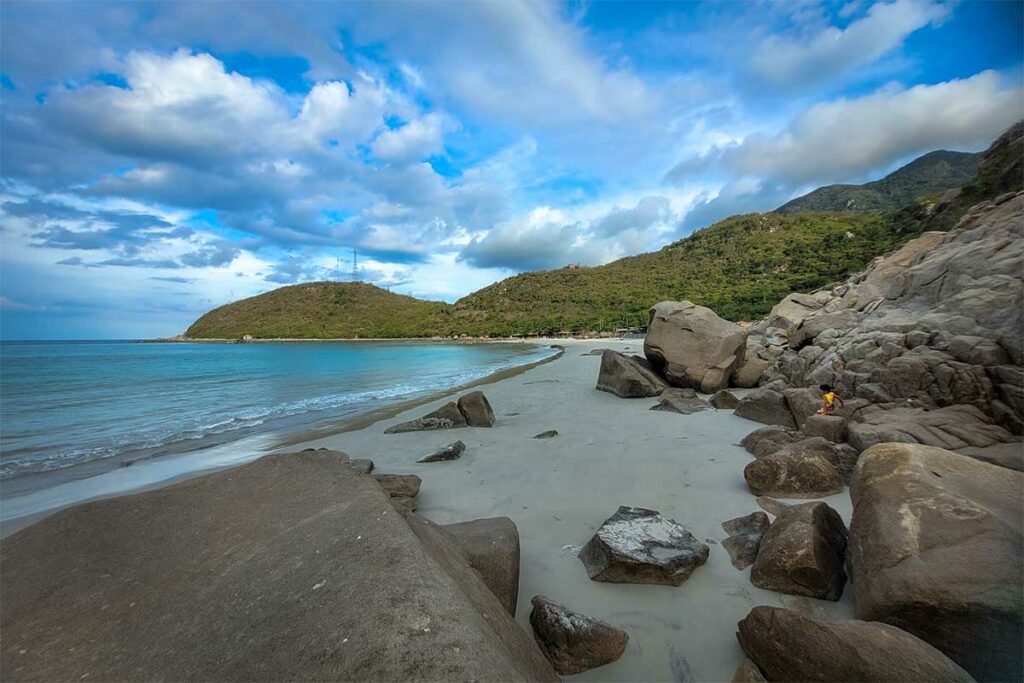 Verlaten strand op het Hon Heo schiereiland in de Van Phong Bay met grote rotsblokken op het witte zand, groen beboste heuvels en lichtblauw water — nauwelijks toeristisch en moeilijk bereikbaar